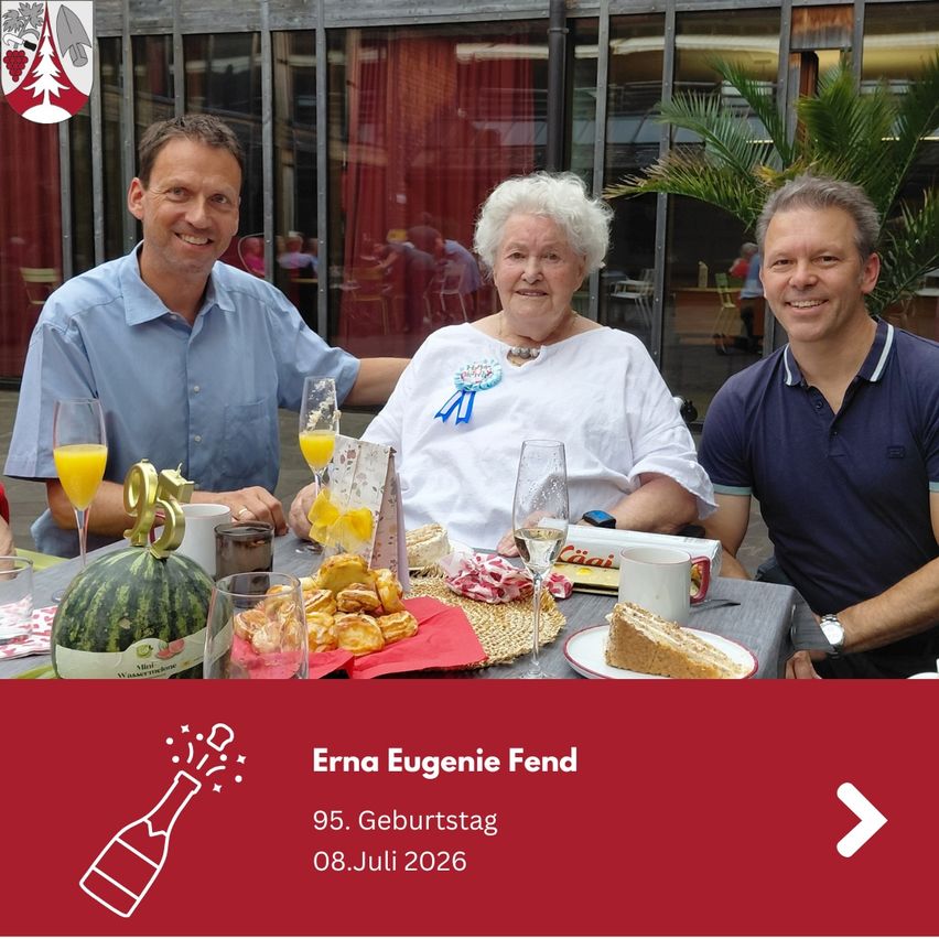 Three people are sitting at a table with food and drinks. A woman in the middle has a blue ribbon on her shirt. The table has a watermelon, a cake, and a cup. The man on the left is holding a glass of orange juice. The man on the right is smiling. The image has a watermark with the text 'Erna Eugenie Fend 95. Geburtstag 08.Juli 2026'.