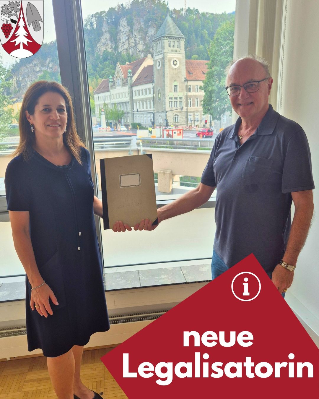 Ein Mann und eine Frau stehen vor einem Fenster und halten eine Mappe. Die Frau lächelt und der Mann trägt eine Brille. Hinter ihnen ist ein Blick auf eine Stadt mit Gebäuden und Bäumen zu sehen.