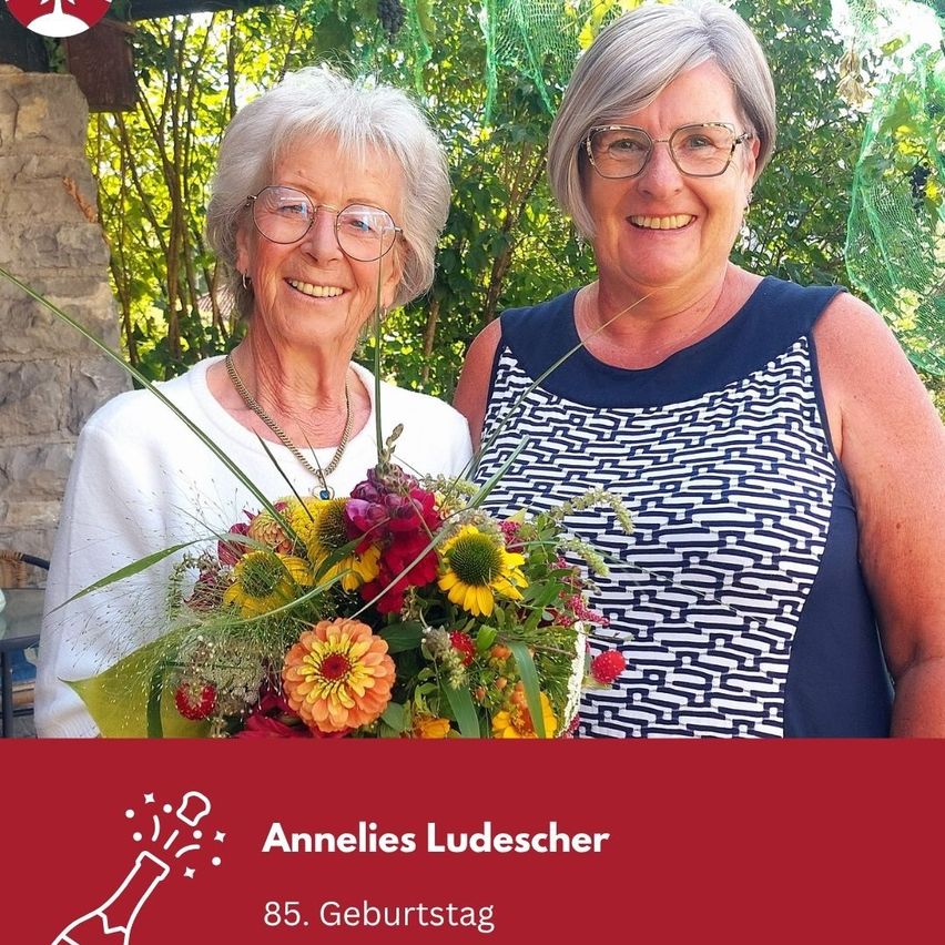 Zwei Frauen lächeln vor einem Garten, eine hält einen Blumenstrauß, mit einer Geburtstagsnachricht für Annelies Ludescher am unteren Rand.