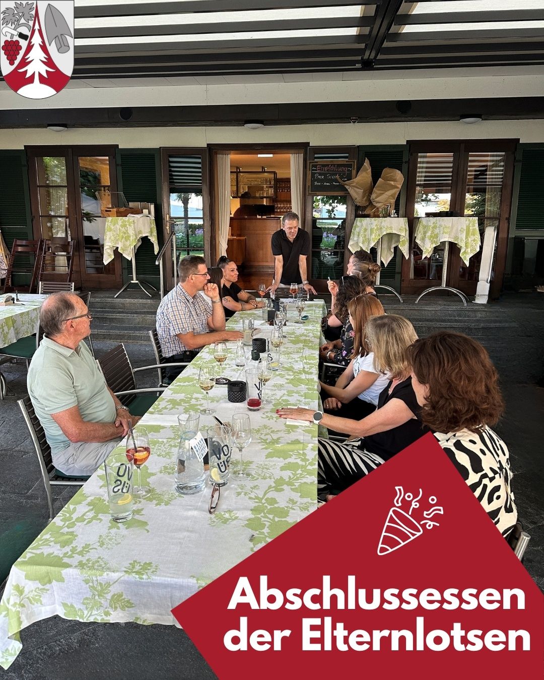 A group of people sit around a long table in an outdoor restaurant. They are looking at a man who is standing and talking. The table has wine glasses, bottles, and plates. There are tables and chairs outside the restaurant.
