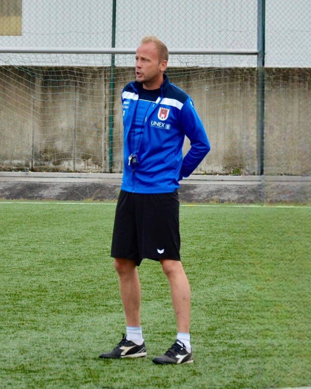 Ein Mann steht auf einem Fußballfeld und trägt eine blaue Jacke mit weißen Abschlüssen und schwarze Shorts. Er ist wahrscheinlich ein Trainer, der das Spiel beobachtet.