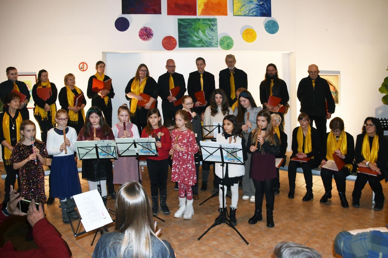 Eine Gruppe von Kindern und Erwachsenen steht in einem Raum und spielt Flöten, während ein Lehrer sie anweist. Bunte Gemälde schmücken die Wand.