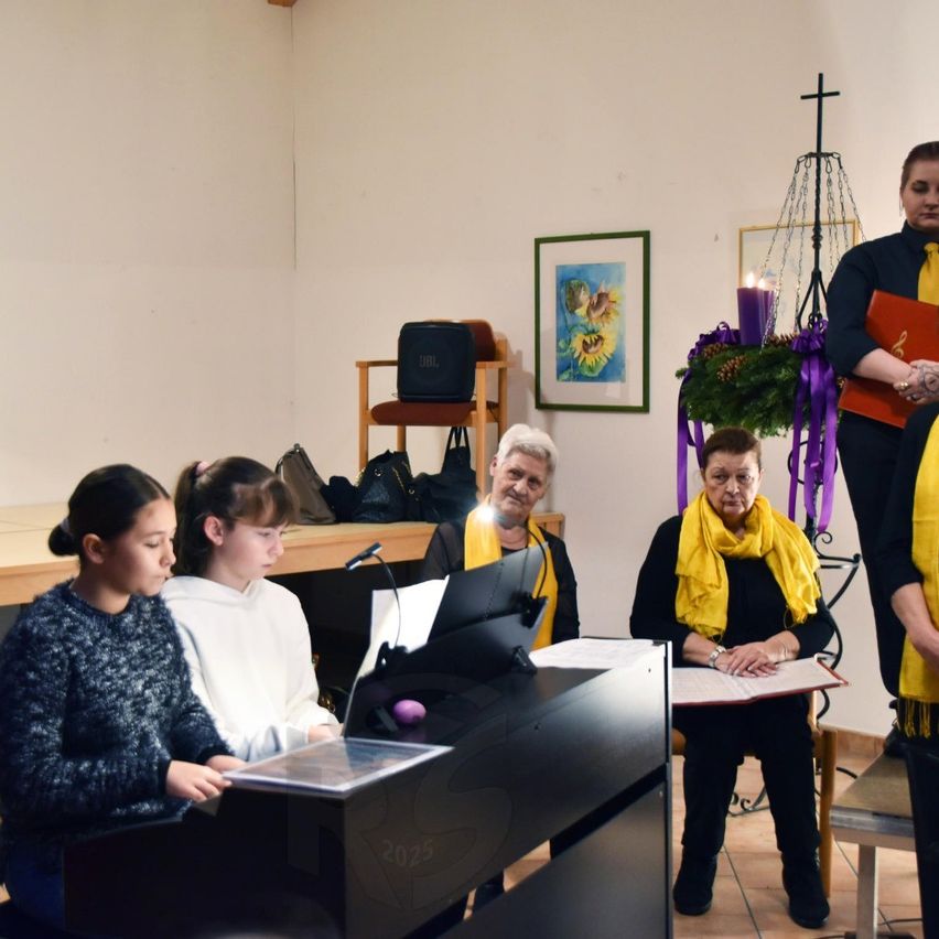 Eine Gruppe von Menschen befindet sich in einem Raum. Zwei Frauen sitzen am Klavier und eine andere steht mit einem Buch. Zwei Frauen sitzen auf dem Boden und lesen Musik. Eine Frau steht mit einem Kreuz und einer Kerze. Ein Bilderrahmen und Taschen stehen auf einem Regal.