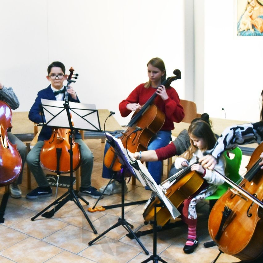 Mehrere Kinder spielen Cello in einem Raum. Ein Mädchen in Rot steht und ein Junge sitzt. Sie sind von Notenständern umgeben.