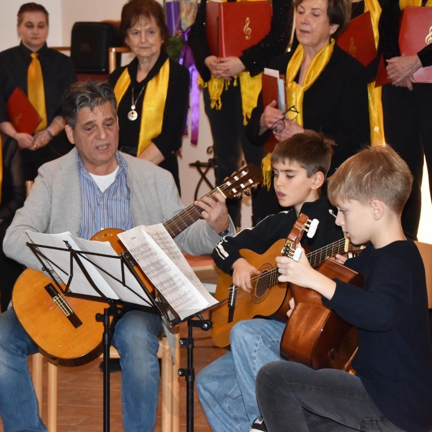 Ein älterer Mann spielt Gitarre, während zwei Jungen in der Nähe sitzen und ebenfalls Gitarre spielen. Dahinter hält ein Chor älterer Frauen Musikbücher. Sie befinden sich alle in einem Raum, möglicherweise während einer Aufführung.