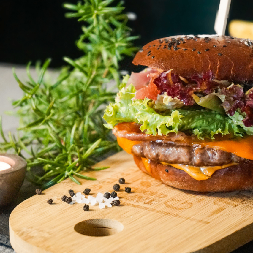 Ein Nahaufnahme eines Burgers mit Salat, Tomate, Käse und Pfeffer auf einem Holzbrett mit Rosmarin und einer Kerze in der Nähe.
