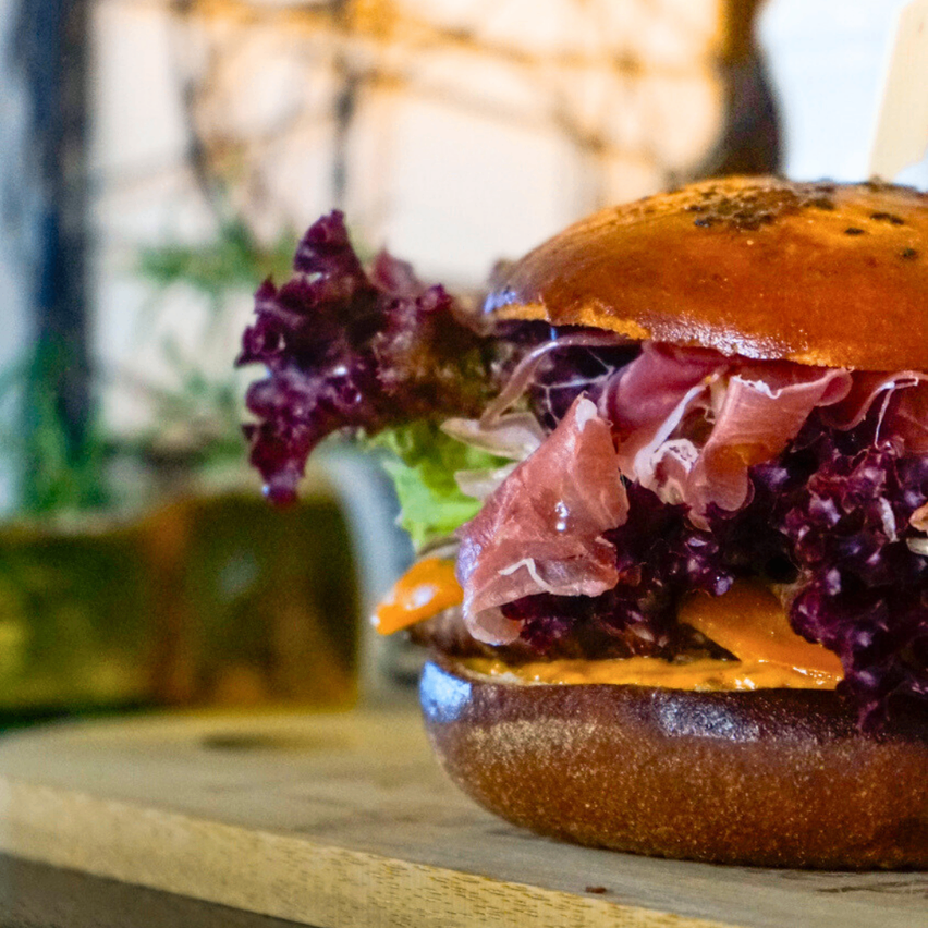 Eine Nahaufnahme eines Gourmet-Burgers mit rotem Salat, geschnittenem Fleisch und Käse auf einer Holzoberfläche. Der Hintergrund ist leicht verschwommen mit einem Hauch von Grün.