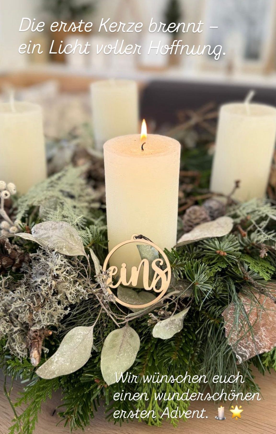 Eine kreisförmige Kranzdekoration mit 'EiNS' in der Mitte, umgeben von weißen Kerzen und Tannengirlanden. Die Kerzen sind entzündet und verbreiten ein warmes Licht.