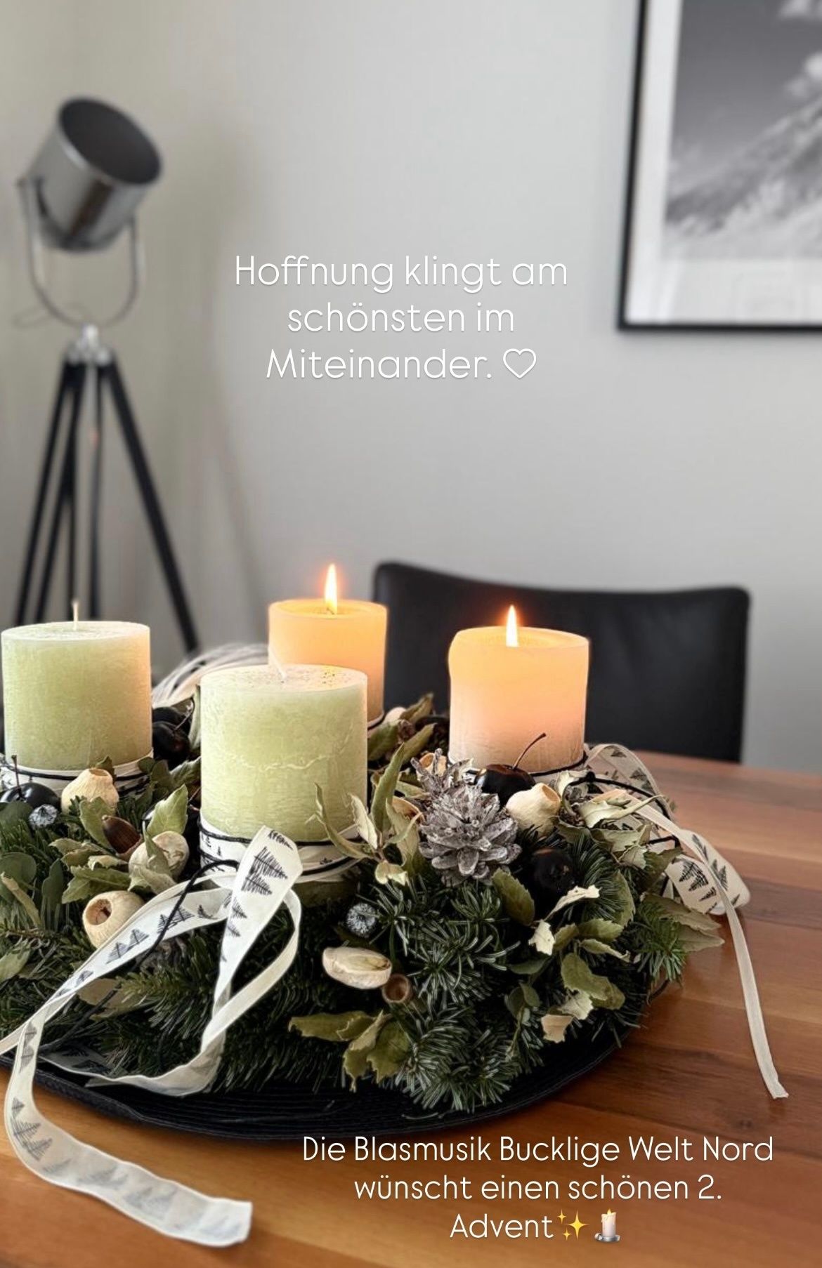 Vier Kerzen sind in einem Kranz auf einem Tisch platziert, umgeben von Grün und Tannenzapfen. Die Kerzen sind angezündet, und ein deutscher Spruch 'Hoffnung klingt am schönsten im Miteinander' steht darüber.