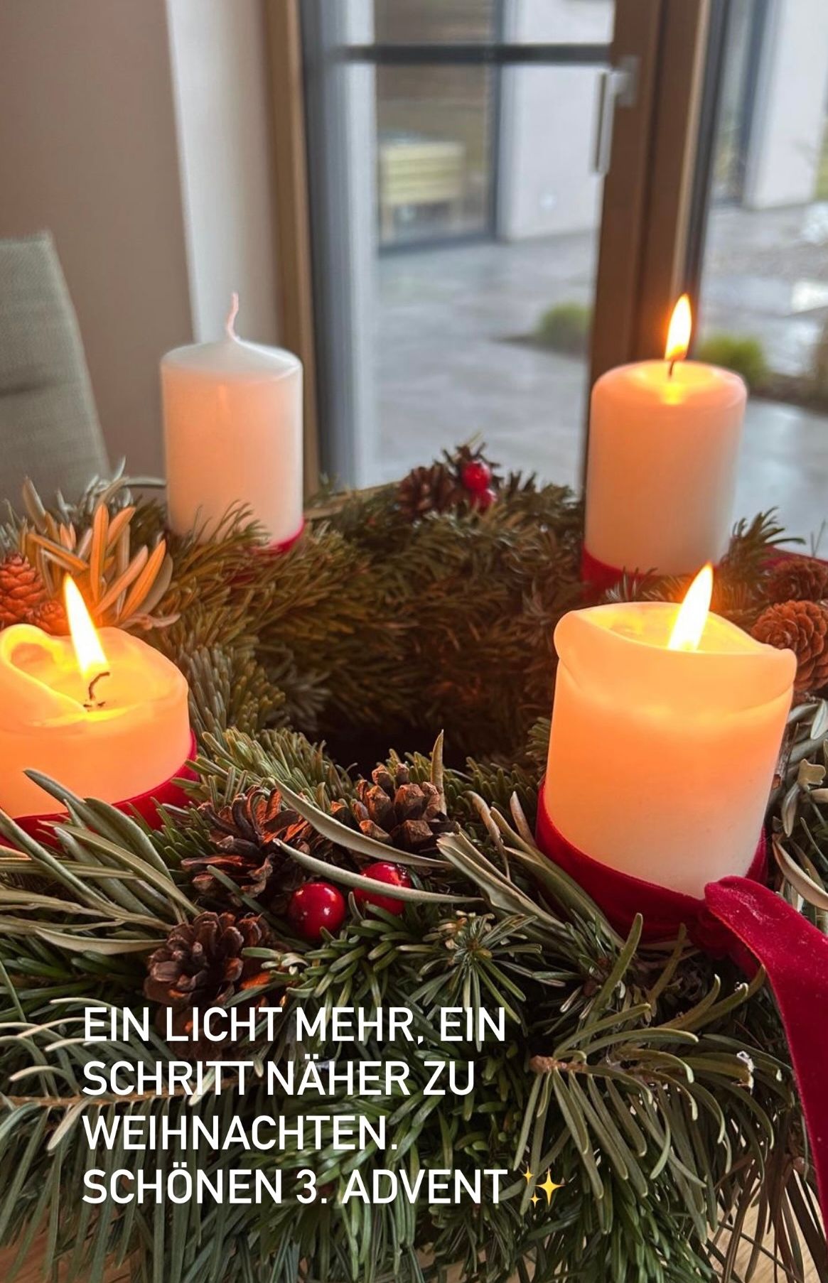 Vier brennende Kerzen mit roten Bändern in einem Kranz aus Kiefernzweigen, verziert mit roten Beeren und Tannenzapfen. Der Kranz steht vor einem Fenster mit Blick auf einen Garten.