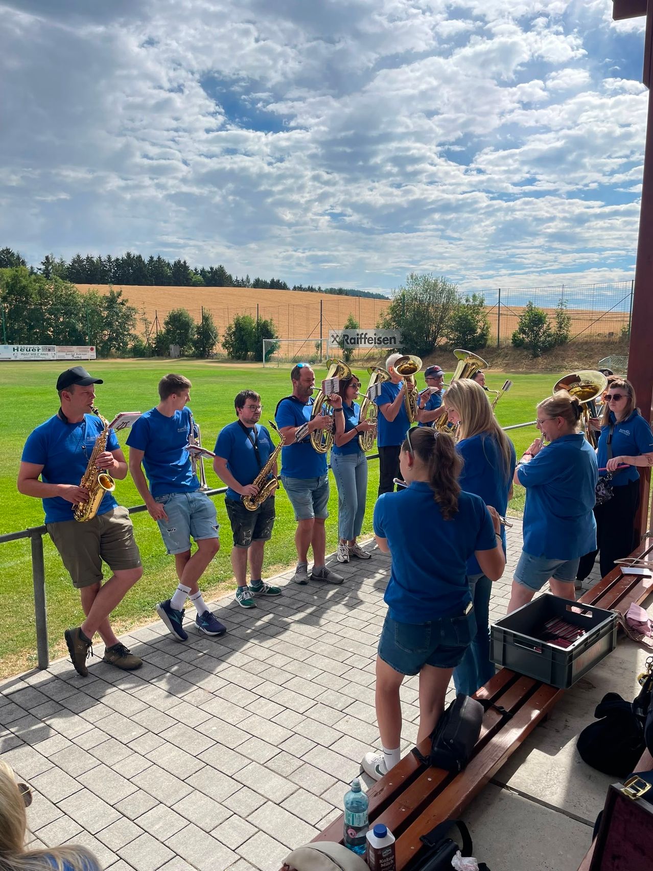 Eine Gruppe von Musikern, die blaue Hemden und Shorts tragen, steht auf einer gefliesten Plattform und hält verschiedene Musikinstrumente in der Hand, möglicherweise in Vorbereitung auf eine Aufführung auf einem Rasenfeld.