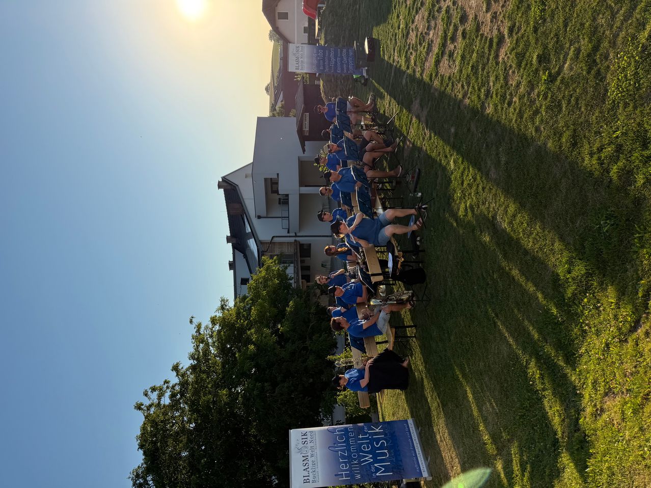 A brass band is seated on a grassy field with a building and banners in the background. The sun is setting in the sky.