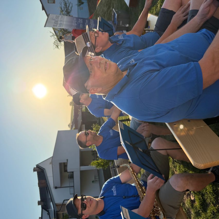 A group of people in blue shirts and hats are gathered on a grassy area, with one person playing the saxophone. The sun is shining brightly in the sky.