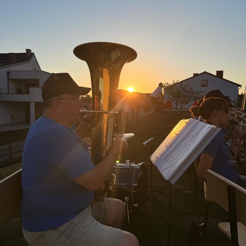 A group of musicians plays instruments outside during sunset. A man in a hat plays a tuba. Another person sits with an open music sheet.