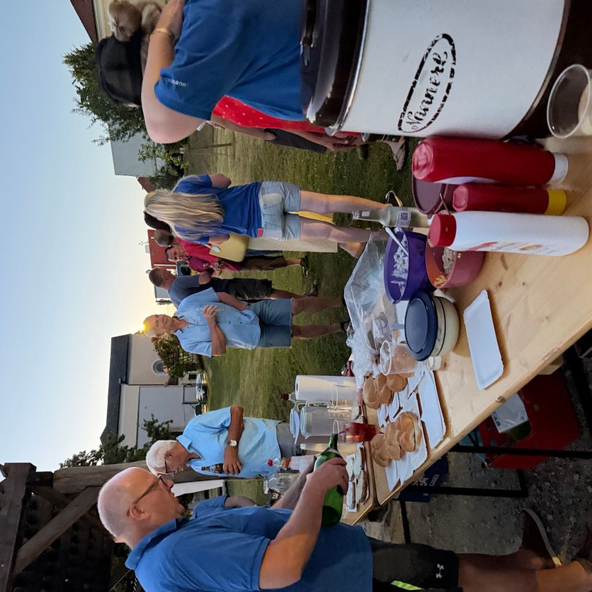 A group of people are gathered around a table outdoors, possibly for a party. Various food items and beverages are on the table. Some people are standing while others are seated. Trees and buildings are visible in the background.