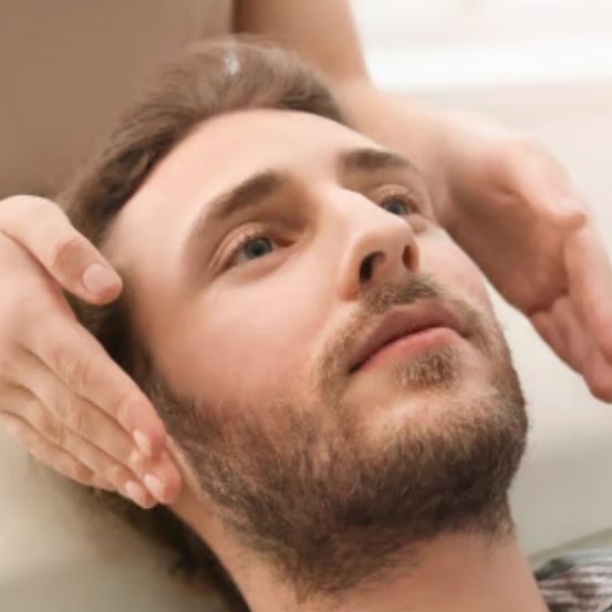 Ein Mann mit Bart liegt auf einer weißen Oberfläche und erhält eine Gesichtsmassage von einer Person, die einen Ring trägt.