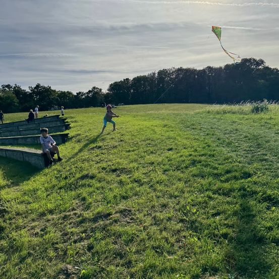 Ein Kind fliegt ein Drachen in einem Grasfeld, während einige Leute auf einer Betonbank im Hintergrund sitzen.