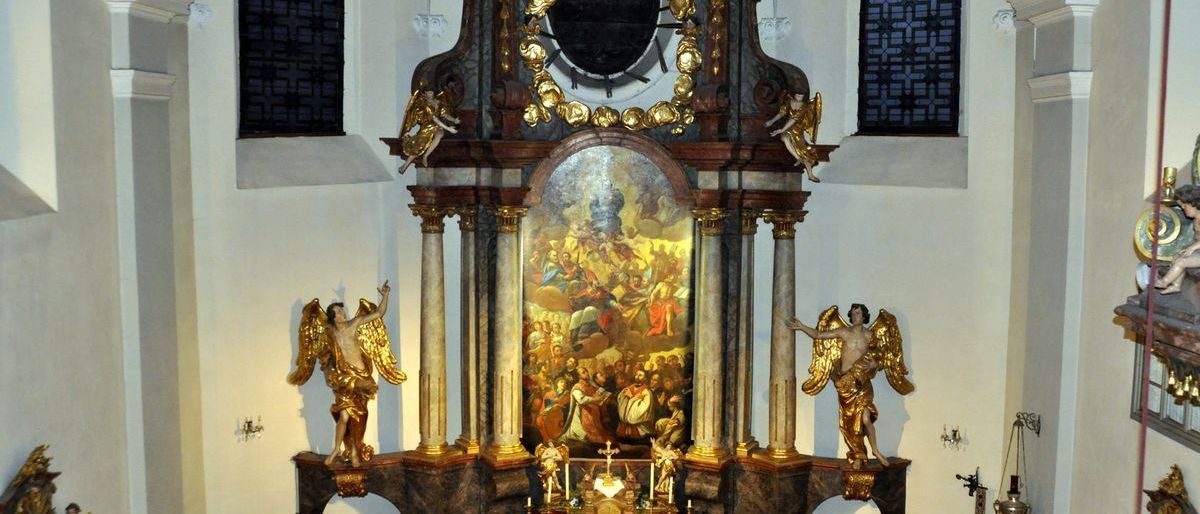Ein kunstvoller Altaraufsatz in einer Kirche mit goldenem Rahmen und zwei Statuen von Engeln. Ein großes Gemälde schmückt den Altar, und zusätzliche dekorative Elemente sind vorhanden.
