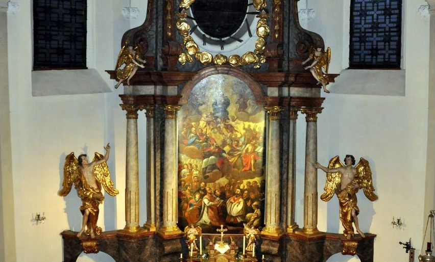 Ein kunstvoller Altaraufsatz in einer Kirche mit goldenem Rahmen und zwei Statuen von Engeln. Ein großes Gemälde schmückt den Altar, und zusätzliche dekorative Elemente sind vorhanden.