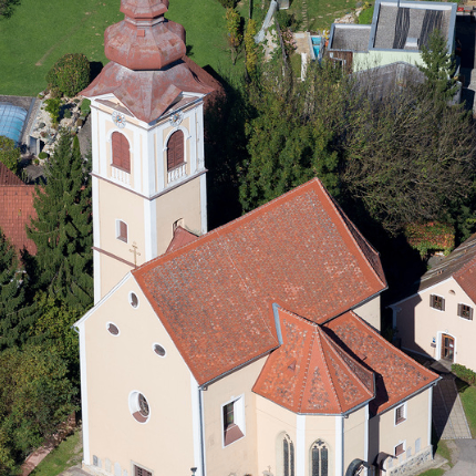 Ein Luftbild einer Kirche mit einem roten Dach und einem hohen Turm, umgeben von Bäumen und Häusern in einer ländlichen Gegend.