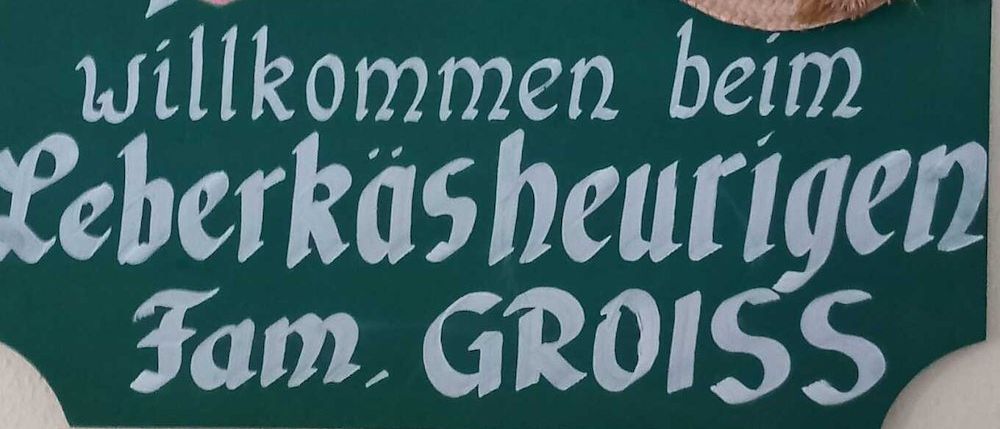 Ein grünes Schild mit weißem Text und einem Herz mit einem Pfeil darin. Es steht 'Willkommen bei Fam. Gross'. Ein Strohhut mit roten Blumen ist auf der rechten Seite des Schildes.