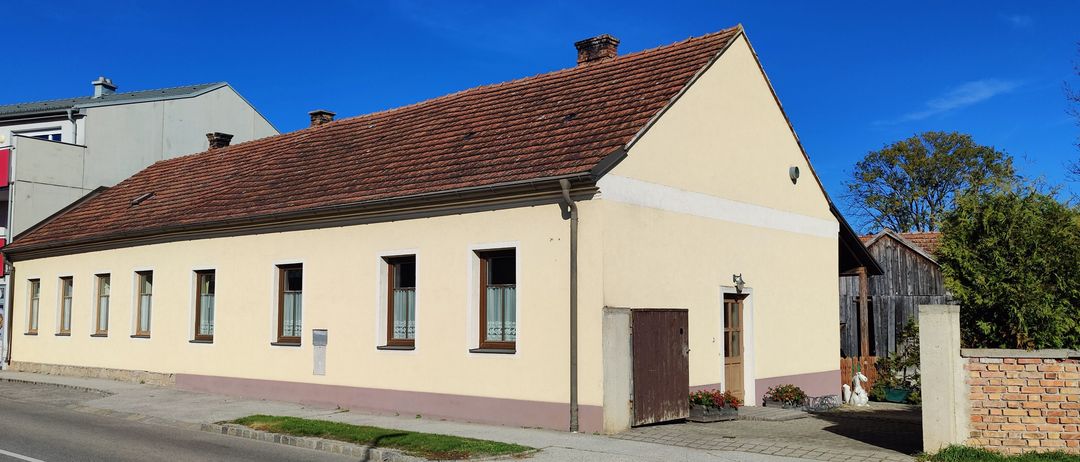 Ein beiges Haus mit rotem Dach und mehreren Fenstern steht an einer Straße mit klarem blauem Himmel.