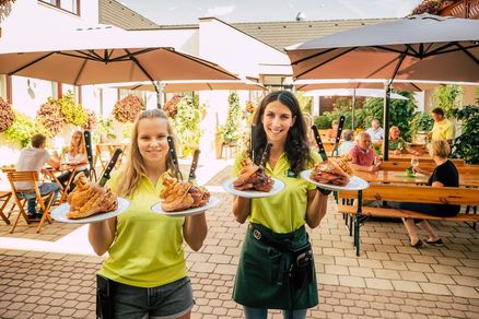 Zwei Frauen in grünen Uniformen halten Teller mit Essen in einem Outdoor-Restaurant. Ein Teller hat frittiertes Essen und der andere gebratenes Fleisch.