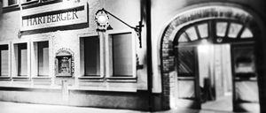 Ein Schwarz-Weiß-Foto eines Gebäudes mit dem Schild Hartberger. Das Gebäude hat mehrere Fenster und eine Lampe an der Wand. Der Eingang ist offen.