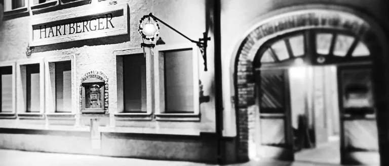 Ein Schwarz-Weiß-Foto eines Gebäudes mit dem Schild Hartberger. Das Gebäude hat mehrere Fenster und eine Lampe an der Wand. Der Eingang ist offen.