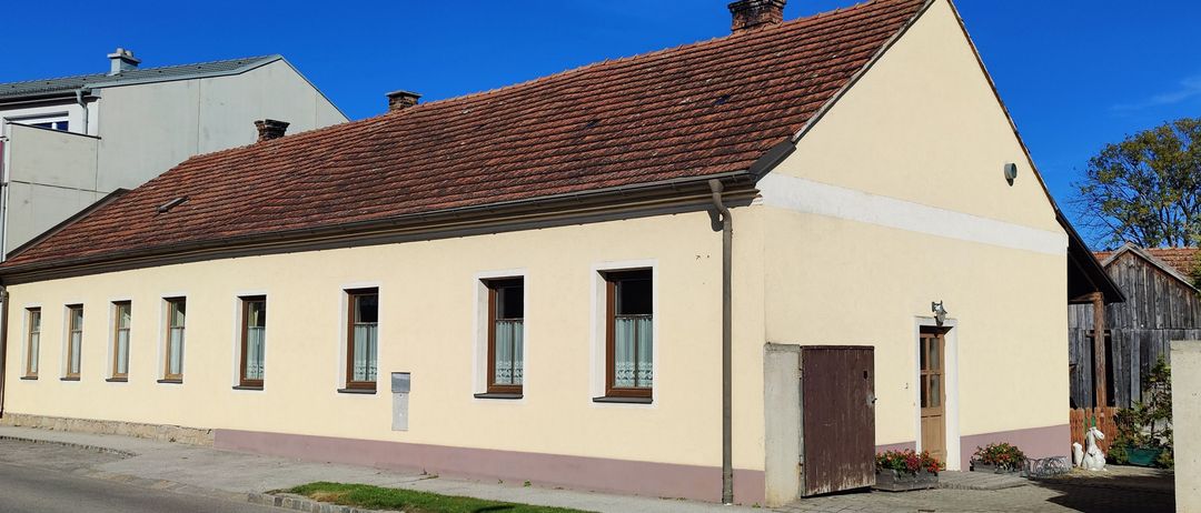 Ein beigefarbenes Haus mit einem braunen Dach und mehreren Fenstern steht an einer ruhigen Straße mit klarem blauem Himmel darüber.