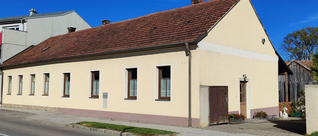 Ein beigefarbenes Haus mit einem braunen Dach und mehreren Fenstern, an einer ruhigen Straße mit einem klaren blauen Himmel darüber.