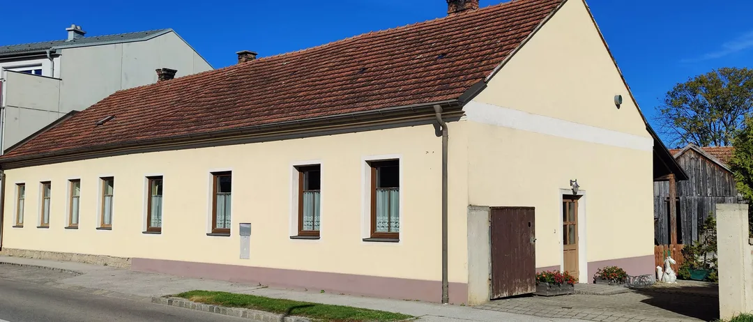 Ein beigefarbenes Haus mit einem braunen Dach und mehreren Fenstern steht an einer Straße unter einem klaren blauen Himmel.