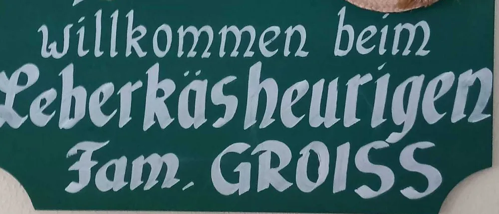 Ein grünes Schild mit weißem Text, das 'Willkommen bei Fam. Gross' und ein Herz mit einem Pfeil durch es zeigt. Ein Strohhut mit Blumen ist auf der rechten Seite des Schildes.