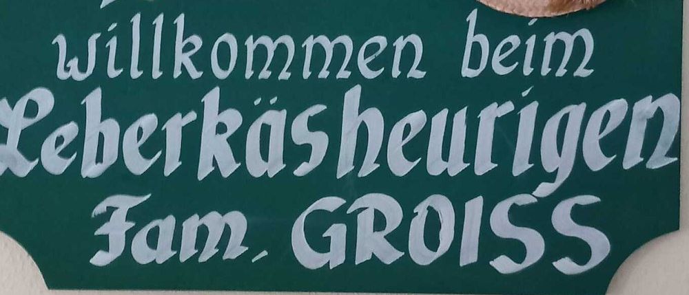 Ein grünes Schild mit weißem Text, das 'Willkommen bei Fam. Gross' und ein Herz mit einem Pfeil durch es zeigt. Ein Strohhut mit Blumen ist auf der rechten Seite des Schildes.