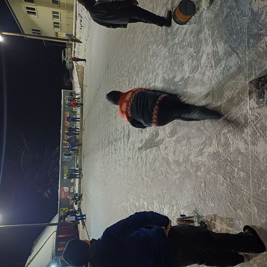 Eine Gruppe von Menschen skat auf einer Eisbahn bei Nacht. Einige tragen Mützen und Turnschuhe. Andere stehen in einer Schlange. Es gibt Straßenlaternen und ein Gebäude in der Nähe.