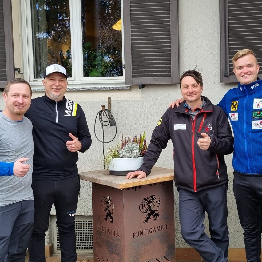 Vier Männer stehen vor einem Gebäude und lächeln für ein Foto. Sie tragen Winterkleidung, wobei einer der Männer einen Daumen hochhält. Ein Topf mit einer Pflanze in Weiß steht auf einem Tisch mit der Aufschrift 'Puntigamer'. Hinter ihnen sind Fenster mit Jalousien zu sehen.