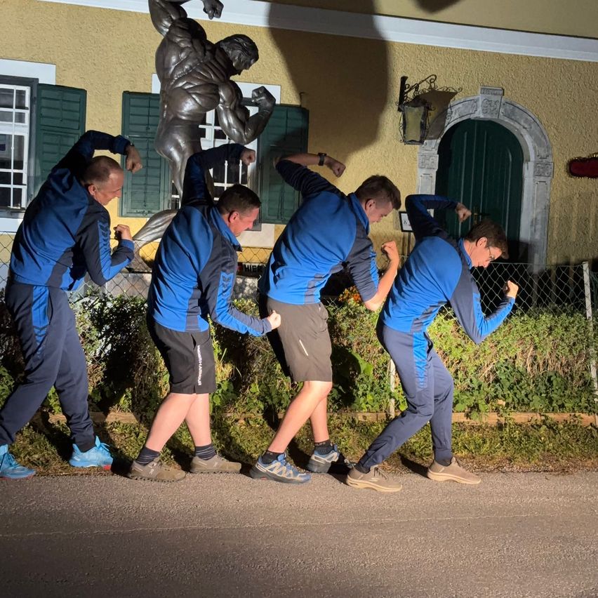 Five men in blue athletic wear perform a synchronized movement in front of a muscular statue on a sidewalk at night.