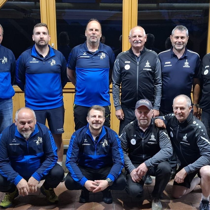 A group of men, likely a sports team, pose for a photo. They wear blue and black jackets with logos. They stand and crouch in front of a wooden wall.