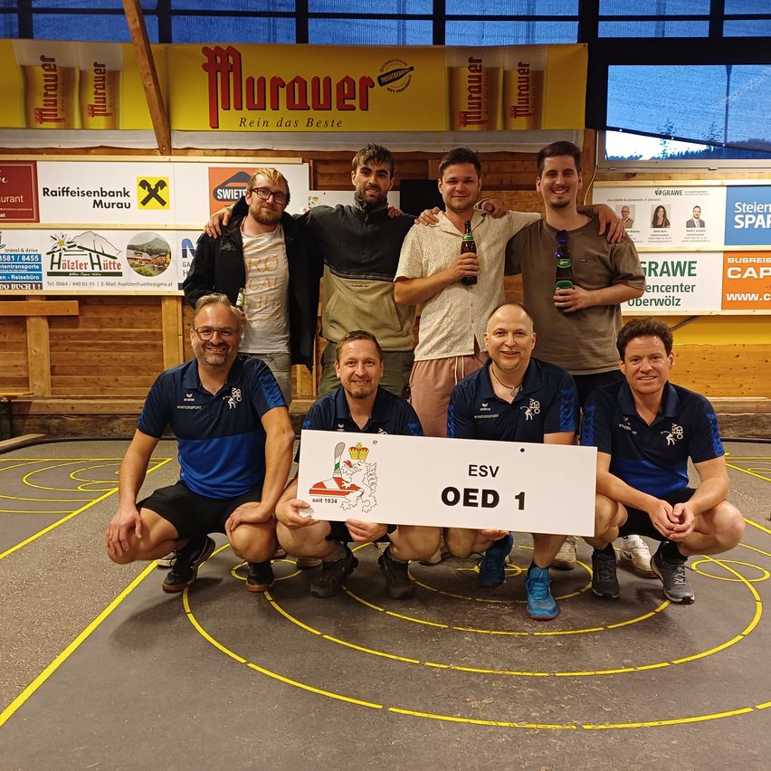 Eine Gruppe von Männern posiert für ein Foto in einem Fitnessstudio, wobei sie ein Schild mit der Aufschrift ESV OED 1 halten. Sie tragen blaue Hemden und einige haben Brillen. Hinter ihnen hängen Banner mit Logos und Texten an der Wand.