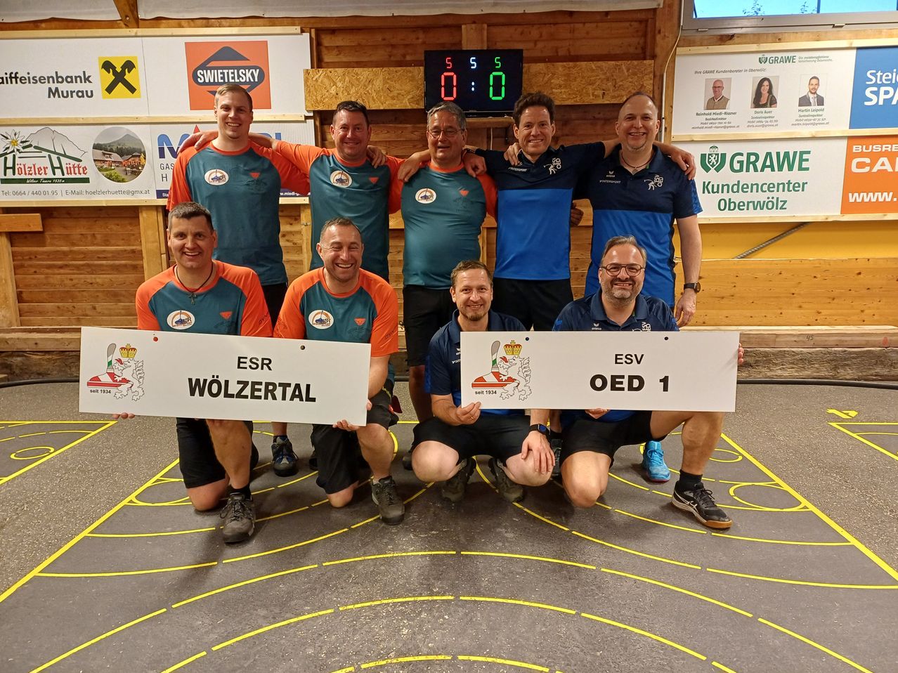 Eine Gruppe von Männern posiert für ein Foto mit einer Anzeigetafel im Hintergrund. Sie tragen Teamtrikots und halten ein Whiteboard mit Text. Die Umgebung scheint ein Sportevent zu sein.