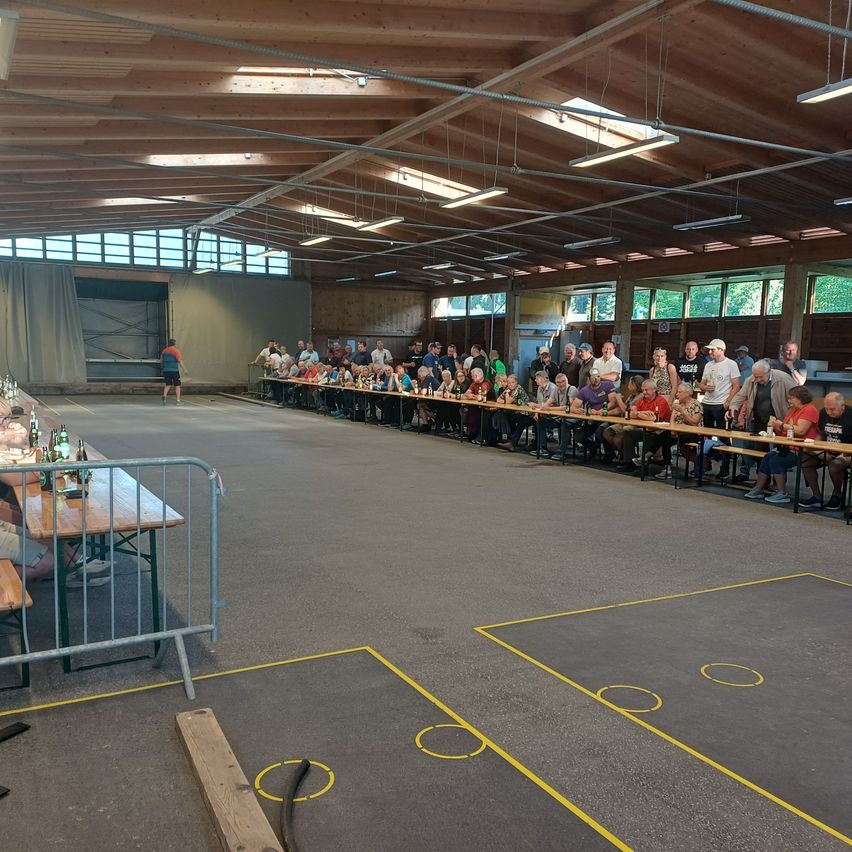 Ein großer Innenbereich mit mehreren Tischen für ein Treffen, mit sitzenden und stehenden Personen, einer Person, die in der Nähe einer Wand geht, und einer Holzbarriere.