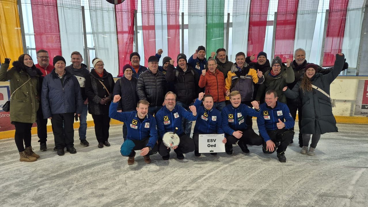 A group of people in blue jackets are posing for a picture at an ice rink. One person is holding a sign that says ESV Oed.