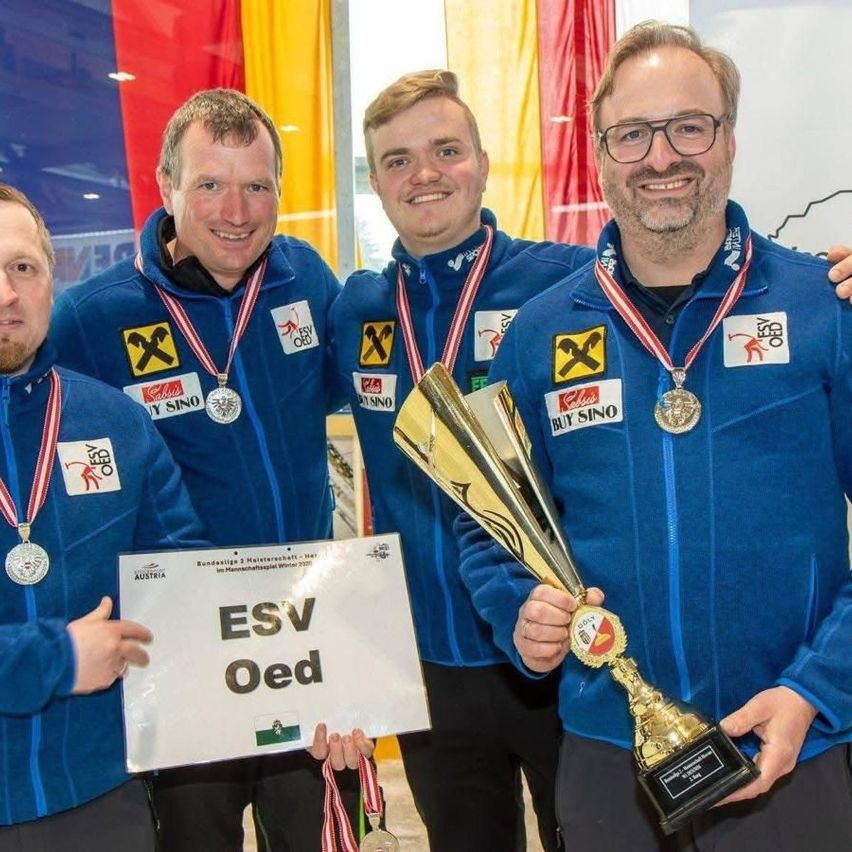 Five men in blue jackets with medals around their necks, holding a trophy and a sign reading ESV Oed. They are standing next to each other and smiling for a photo. Behind them, there is a colorful background with flags.