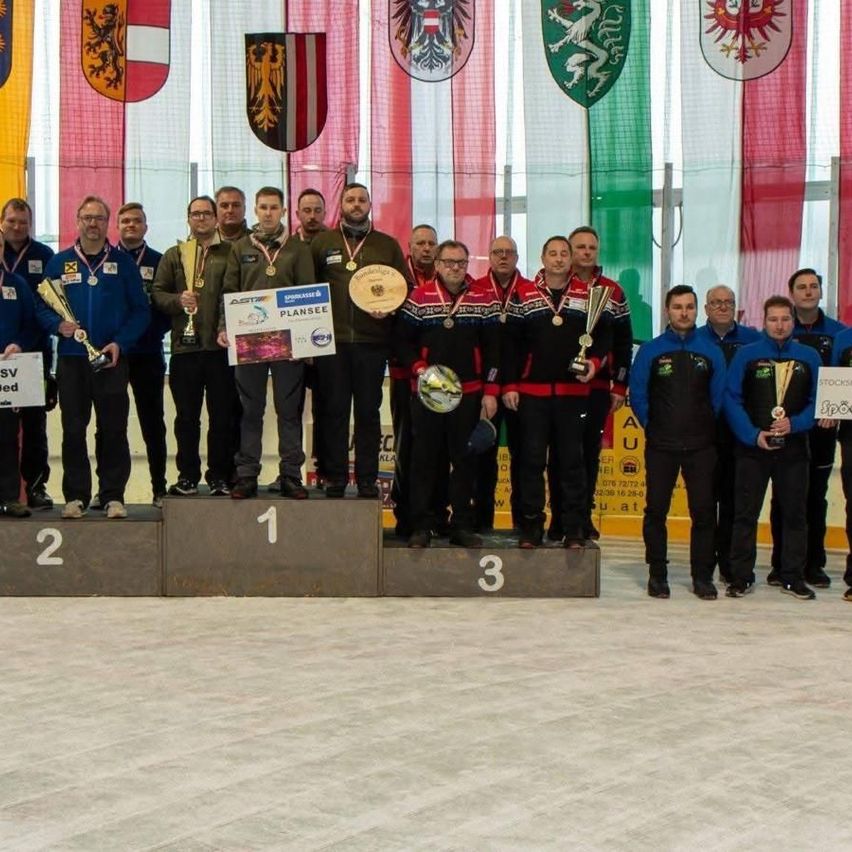 A group of men stand on a podium, holding trophies and medals. They are likely athletes participating in a sports event. Behind them, flags of different countries are displayed.