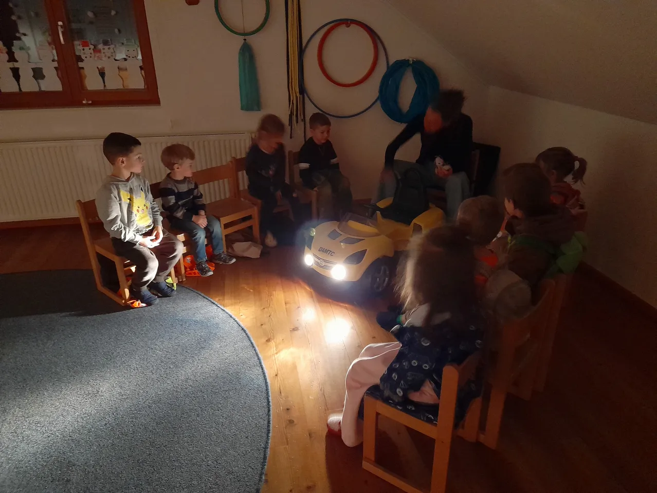 Eine Gruppe von Kindern sitzt auf Holzstühlen in einem Raum und hört einem Mann zu. Ein Spielzeugauto steht vor ihnen.