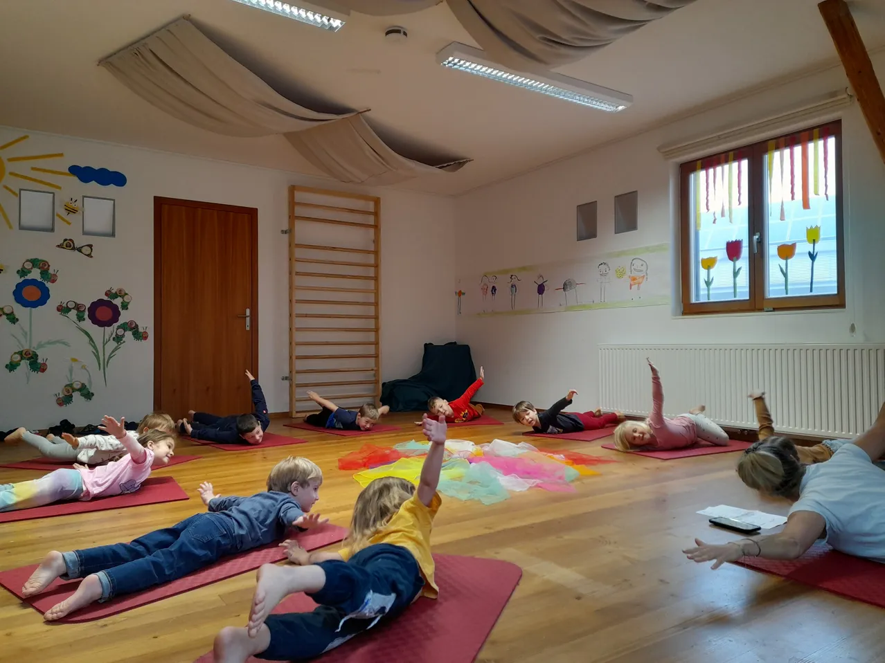 Kinder in einem Klassenzimmer praktizieren Yoga auf dem Boden mit verschiedenen Yoga-Matten und bunten Objekten auf dem Boden.