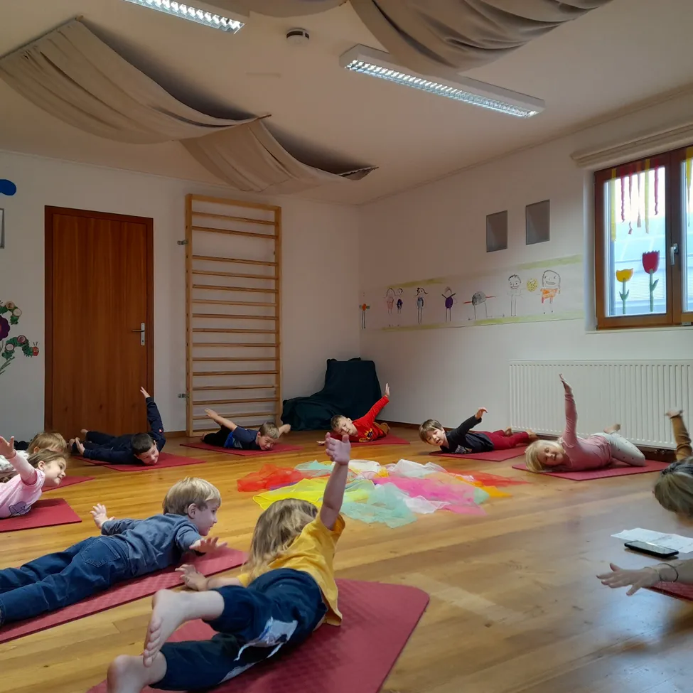 Kinder in einem Klassenzimmer praktizieren Yoga auf dem Boden mit verschiedenen Yoga-Matten und bunten Objekten auf dem Boden.