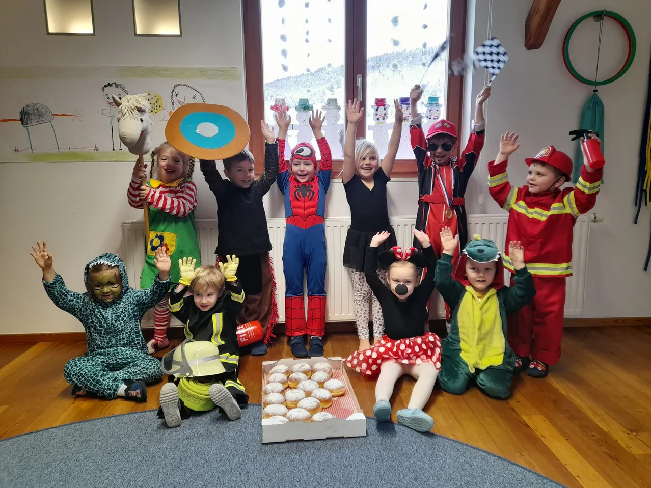 Eine Gruppe von Kindern in Kostümen posiert für ein Foto. Einige Kostüme sind Superhelden, Feuerwehrleute und ein Krokodil. Auf dem Boden steht eine Schachtel Donuts.