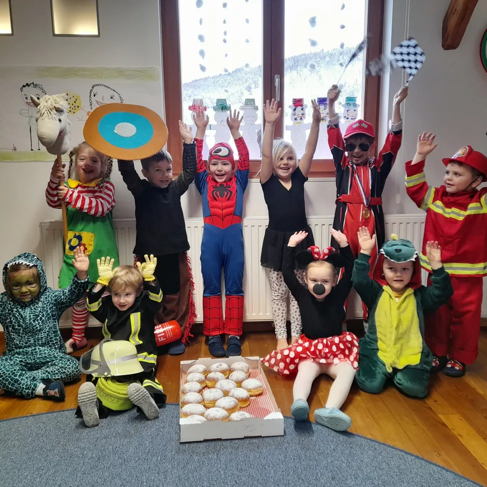 Eine Gruppe von Kindern in Kostümen posiert für ein Foto. Einige Kostüme sind Superhelden, Feuerwehrleute und ein Krokodil. Auf dem Boden steht eine Schachtel Donuts.
