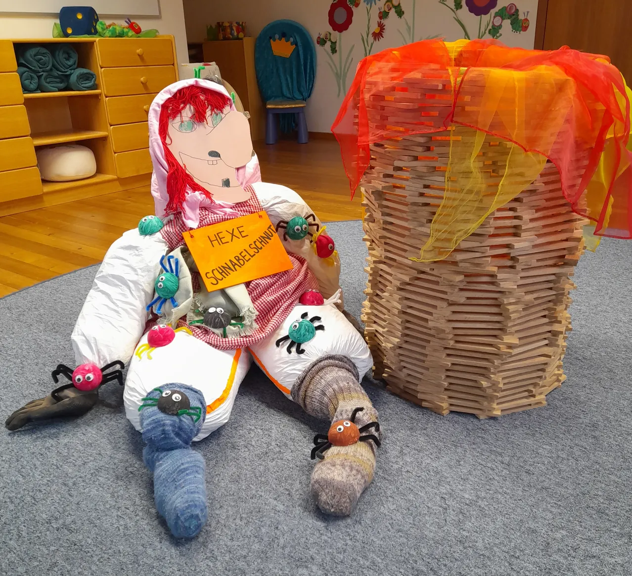 Eine Puppe mit roten Haaren sitzt neben einer Holzkonstruktion mit bunter Abdeckung auf dem Boden. Die Puppe hat ein Schild mit der Aufschrift 'Hexe Schnabbelschnautz'. Spinnen sitzen auf der Puppe und der Holzkonstruktion.